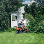 Husqvarna front deck ride-on mower cutting a well-maintained lawn with shrubs in the background on a sunny day